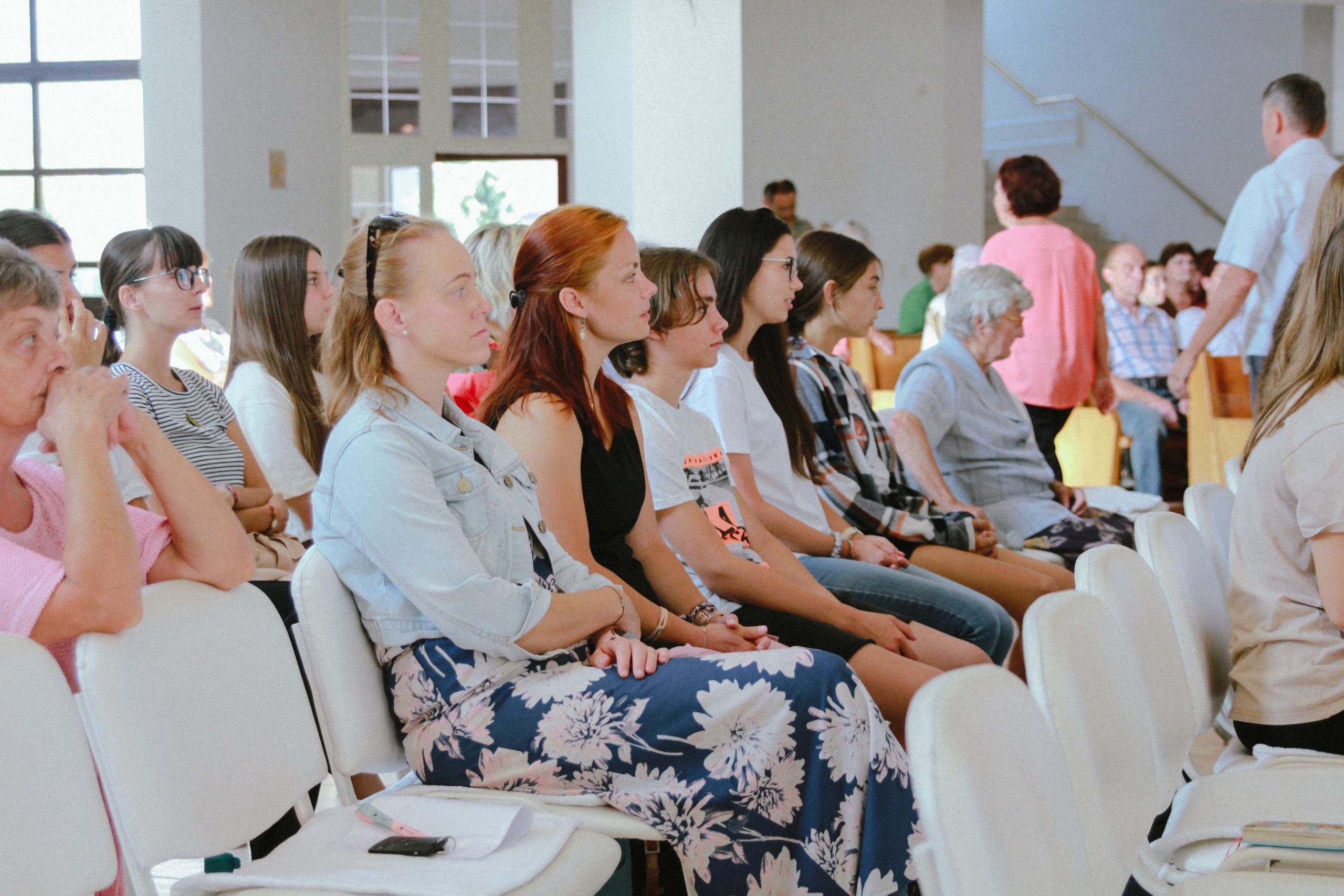 A diverse group of people sitting attentively during an indoor meeting or event, captured in a spacious room.